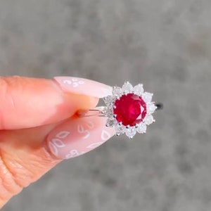 May include: A silver ring featuring a large red ruby surrounded by smaller white diamonds. The ring is set in a flower-shaped design.