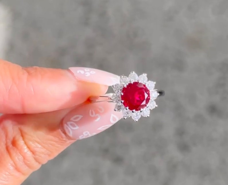 May include: A silver ring featuring a large red ruby surrounded by smaller white diamonds. The ring is set in a flower-shaped design.