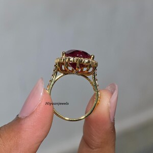 Pu&ograve; includere: Un anello d'oro con una grande pietra preziosa rosso scuro a taglio ovale, circondata da pietre chiare pi&ugrave; piccole. La fascia dell'anello &egrave; anch'essa ornata da piccole pietre chiare. Il testo "Hiyaanjewels" &egrave; visibile sull'anello.