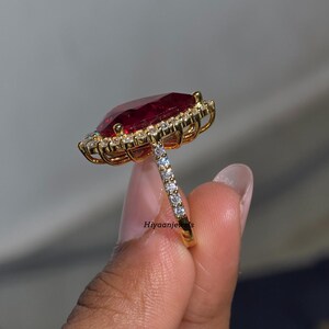 Pu&ograve; includere: Un anello d'oro con una grande pietra preziosa rosso scuro rettangolare, circondata da pietre chiare pi&ugrave; piccole. La fascia dell'anello &egrave; anch'essa ornata da piccole pietre chiare. L'anello &egrave; mostrato su uno sfondo morbido e neutro.