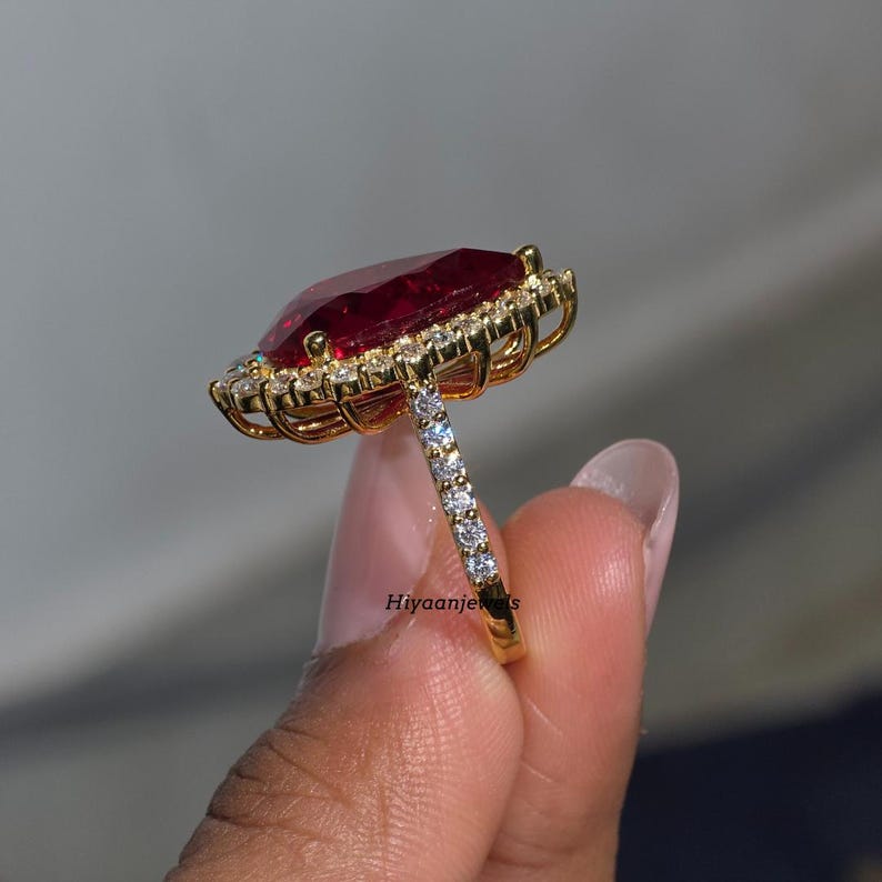 Pu&ograve; includere: Un anello d'oro con una grande pietra preziosa rosso scuro rettangolare, circondata da pietre chiare pi&ugrave; piccole. La fascia dell'anello &egrave; anch'essa ornata da piccole pietre chiare. L'anello &egrave; mostrato su uno sfondo morbido e neutro.