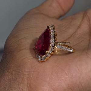 Pu&ograve; includere: Un anello d'oro con una grande gemma rossa a forma di pera, circondata da pietre bianche pi&ugrave; piccole. La fascia &egrave; anch'essa ornata di pietre bianche. Il testo "Hiyaanjewels" &egrave; visibile sull'anello.