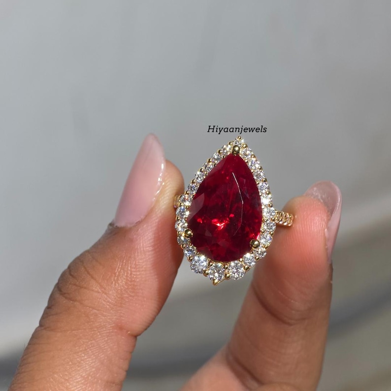 Pu&ograve; includere: Un anello d'oro con una grande pietra preziosa rossa a forma di pera, circondata da pietre preziose pi&ugrave; piccole e trasparenti. L'anello &egrave; tenuto su uno sfondo neutro. Il testo "Hiyaanjewels" &egrave; visibile.