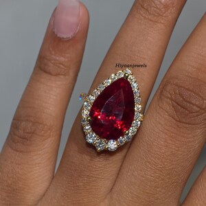 Pu&ograve; includere: Un anello d'oro con una grande gemma rosso scuro a forma di pera, circondata da un alone di gemme pi&ugrave; piccole e chiare. La fascia dell'anello &egrave; anch'essa dorata. Il testo "Hiyaanjewels" &egrave; visibile.