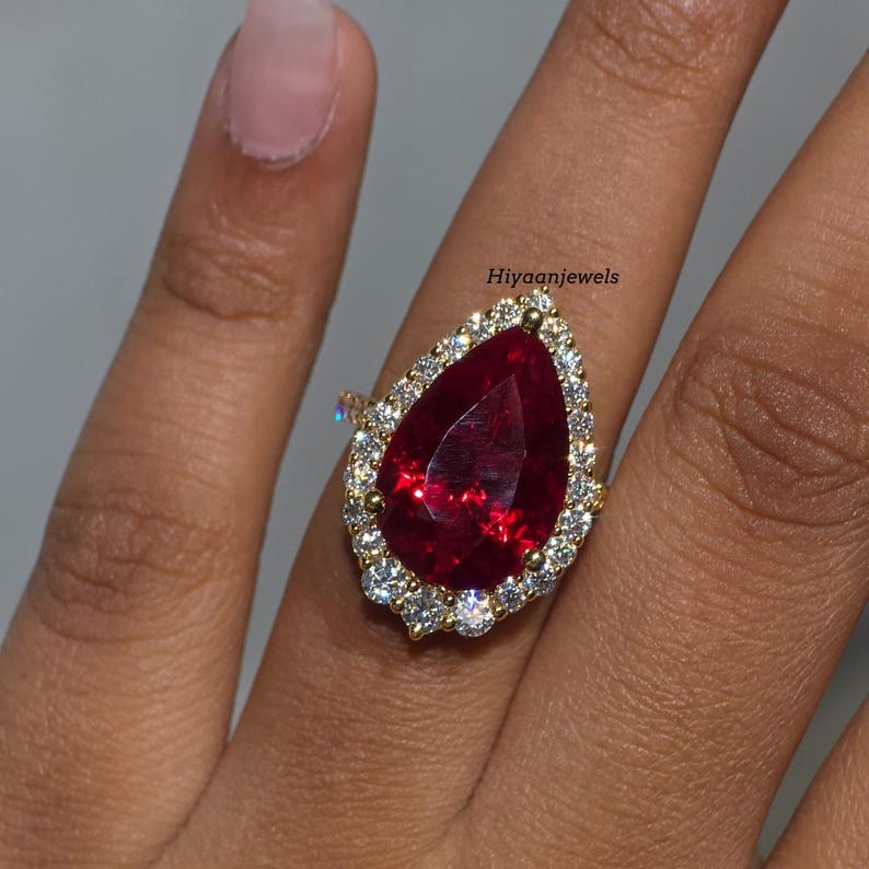Pu&ograve; includere: Un anello d'oro con una grande gemma rosso scuro a forma di pera, circondata da un alone di gemme pi&ugrave; piccole e chiare. La fascia dell'anello &egrave; anch'essa dorata. Il testo "Hiyaanjewels" &egrave; visibile.