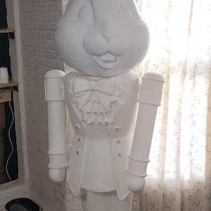 Puede incluir: Una gran estatua blanca de un conejo vestido con un traje formal, que incluye pajarita y botas. La estatua tiene orejas grandes y una cara sonriente. Se encuentra sobre una base redonda.