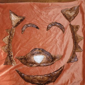 Può includere: Un cuscino arancione con un design a forma di viso realizzato con pezzi marroni strutturati. Il viso comprende due sopracciglia curve, una bocca sorridente e un baffo con un centro bianco. Due pezzi appuntiti incorniciano il viso.