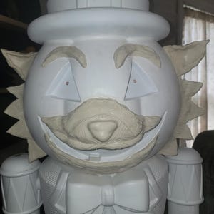 Può includere: Una grande statuetta decorativa schiaccianoci bianca. La statuetta ha un cappello, un volto sorridente con occhi triangolari e un papillon. I dettagli sono scolpiti e il design generale è stravagante e festoso.