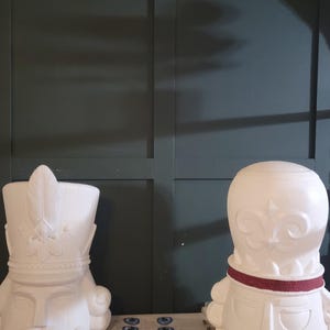 May include: Two white, decorative sculptures with detailed facial features and headwear. One has a crown with a feather, the other a red band. A sheet of paper with eye designs is between them. The background is a dark green wall.