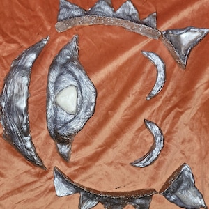 Può includere: Una collezione di pezzi d'arte astratta color argento disposti su un tessuto arancione. I pezzi includono forme curve, una forma centrale a forma di occhio con una pietra bianca e bordi frastagliati, forse per un progetto di scultura o artigianato.