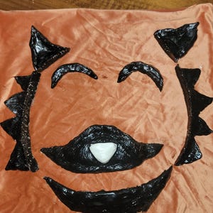 Può includere: Un cuscino arancione con un design a forma di viso a fumetto nero. Il viso presenta sopracciglia nere, baffi e una bocca sorridente. Un triangolo bianco è al centro della bocca.