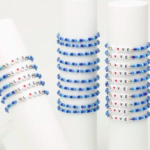 Pode incluir: Uma coleção de pulseiras elásticas feitas de pequenas contas de vidro azul e azul claro. Cada pulseira apresenta contas de letras brancas que formam a palavra LOVE, com um pequeno coração vermelho substituindo a letra O e contas prateadas.