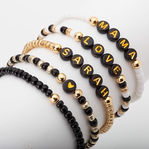 May include: A collection of beaded bracelets in black, gold, and white. Some bracelets spell out "MAMA", "LOVE", and "SARAH" with gold letters on black beads. Other bracelets feature solid gold and black beads.