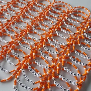 Pode incluir: Uma coleção de pulseiras elásticas feitas de pequenas contas laranja e contas brancas com letras que formam a palavra TODAY. Cada pulseira apresenta uma única conta prateada metálica intercalada entre as contas laranja e brancas.