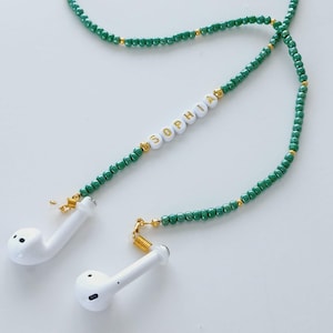 May include: Earbud strap with green beads and gold accents. White beads spell out the name "SOPHIA". The strap is connected to white earbuds.