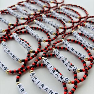 Pode incluir: Uma coleção de pulseiras de contas. Cada pulseira apresenta o nome "ARIRANG" escrito com contas de letras brancas. As pulseiras têm contas vermelhas e pretas, com detalhes dourados. As pulseiras estão espalhadas numa superfície clara.