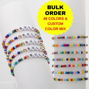 Könnte beinhalten: Mehrere elastische Armbänder mit kleinen, bunten Glasperlen und goldfarbenen Metallzwischenstücken. Fünf Armbänder zeigen weiße Buchstabenperlen, die das Wort BULK bilden. Eine gelbe Grafik zeigt den Text BULK ORDER 48 COLORS & CUSTOM COLOR MIX.