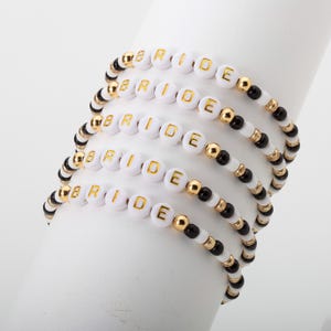 May include: Five beaded bracelets spelling "BRIDE" with white letter beads and gold letters. The bracelets have a mix of white, black, and gold beads. The bracelets are stacked on a white cylindrical object.