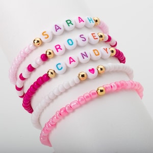 Könnte beinhalten: Fünf bunte Perlenarmbänder mit weißen Perlen und goldenen Akzenten. Die Armbänder buchstabieren die Namen "SARAH", "ROSE", "CANDY" und beinhalten einen Herzanhänger.