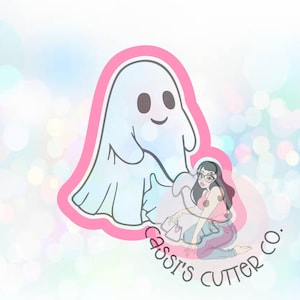 May include: A white ghost with a friendly face and a pink outline. The ghost is holding a small pink bag with a white heart on it. The image is on a light blue background with a pink and white logo that says "Cassi's Cutter Co."