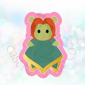 May include: A pink cookie cutter shaped like a green alien with red hair wearing a yellow and green dress.