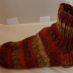 Op de afbeelding: Gestreepte gebreide sok in tinten van bruin, oranje en groen. De sok is bovenaan gevouwen, waardoor het breipatroon zichtbaar wordt.