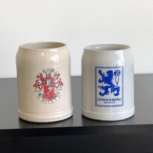 May include: Two ceramic beer steins. One is beige with a red and white crest and the word "BECK'S". The other is white with a blue lion emblem and the words "LOWENBRAU MUNICH".