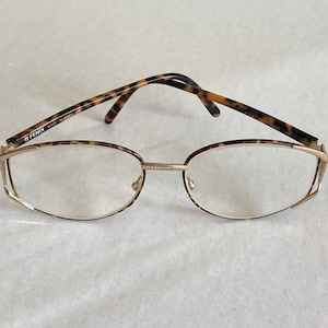 May include: Gold-rimmed eyeglasses with a tortoiseshell pattern on the temples. The glasses have a delicate, oval shape and a thin gold bridge. The brand name "FENDI" is visible on the temple. These stylish glasses are a fashion accessory.