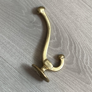 May include: A polished brass wall hook featuring a curved, elongated upper arm and a shorter, lower hook, both ending in rounded ball finials. The base is oval-shaped with two visible screw holes for mounting, showcasing a smooth, metallic finish.