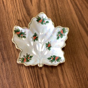 May include: A decorative, leaf-shaped dish with a pearlescent white finish. The dish is adorned with green holly leaves and red berries, and has a gold-colored trim. The dish is a festive holiday accent.