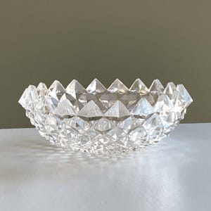 May include: A clear, faceted glass bowl with a diamond-like pattern. The bowl has a scalloped rim with triangular points. The bowl is likely used for decorative purposes or serving small items.