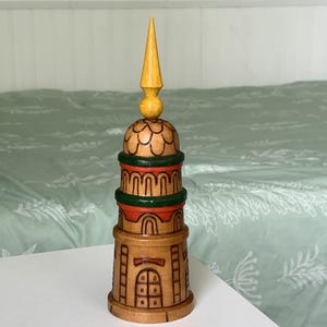 May include: A cylindrical wooden tower figurine featuring a pointed yellow spire top, a scalloped dome section, two green and orange horizontal bands, and a base with carved rectangular window details and vertical lines in a natural wood finish.