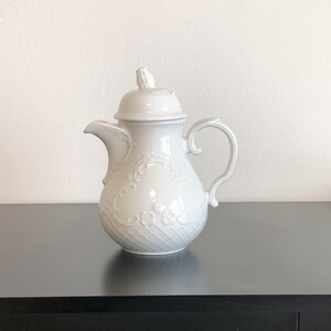 May include: A white ceramic teapot with a lid, spout, and handle. The teapot features embossed floral and swirl designs. The teapot is sitting on a dark surface against a white wall.