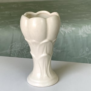 May include: A cream-colored ceramic vase featuring a sculpted tulip-like design with overlapping petal textures. The vessel has a flared rim, a tapered body, and a circular base, showcasing a subtle crackle glaze finish across its entire surface.