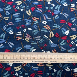 May include: A dark blue fabric featuring a repeating pattern of dragonflies in shades of red, light blue, white, and tan. A wooden ruler is positioned at the bottom, showing a scale in centimetres and millimetres for size reference.