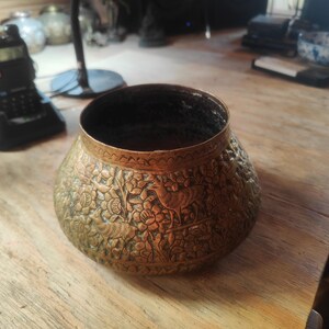 May include: A round, antique-style brass bowl featuring intricate, embossed relief carvings of deer and floral patterns across its exterior surface. The vessel has a wide, open top with a smooth, dark interior and a weathered, aged metallic finish.