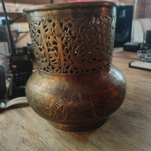 May include: A cylindrical brass vessel featuring intricate openwork filigree patterns on the upper section and detailed engraved Arabic calligraphy and geometric motifs on the rounded lower body, showcasing a metallic gold-toned finish throughout.