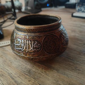 May include: A round, metallic bowl featuring a dark bronze finish with intricate, raised silver-toned Arabic calligraphy and detailed geometric patterns embossed across the exterior surface. The vessel has a wide opening and a curved, bulbous shape.