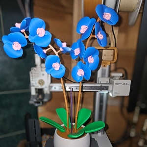 May include: A 3D printed orchid plant with blue and pink flowers in a white pot with brown soil. The plant is sitting on a gray platform with the word "Hotends" printed on it.