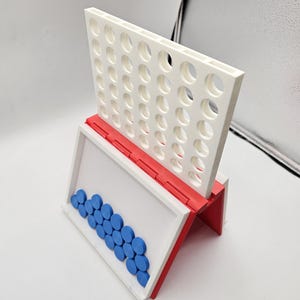 May include: A white and red tabletop game with a grid of holes and blue game pieces. The game is designed to be played by two players, with the goal of connecting four of their colored pieces in a row.