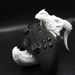 May include: A black video game controller is held in the mouth of a white 3D printed dinosaur skull.