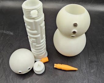 May include: A white snowman kit with three pieces, a carrot nose, and two black buttons for eyes. The snowman is made of plastic and is ready to be assembled.