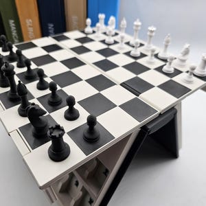 Op de afbeelding: Een draagbare schaakset met zwarte en witte stukken op een dambord. Het bord is wit met zwarte vierkanten en de stukken zijn in hun startposities geplaatst. De schaakset staat op een wit oppervlak.