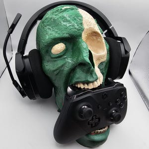May include: A green and bone-colored skull-shaped gaming headset stand. The skull holds a black Xbox controller in its mouth and has a black gaming headset resting on its head. The stand is a unique accessory for gamers.