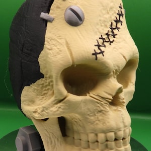 May include: A beige and black skull sculpture with stitched seams and a screw in the forehead. The skull is sitting on a grey base.