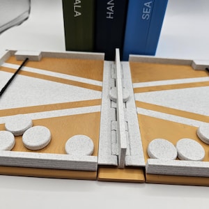 May include: A backgammon board with a white and gold color scheme. The board is open, revealing the playing surface with white and gold triangles. White game pieces are placed on the board. The board is hinged in the middle.