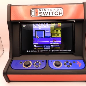 May include: A Nintendo Switch console housed in a miniature arcade cabinet. The cabinet is red and black with a blue control panel. The screen displays a retro video game. The text "NINTENDO SWITCH" is visible on the top.