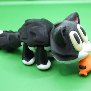 May include: Black and white cat toy with a black and orange fish in its mouth. The cat toy is made of plastic and has a cartoonish design.