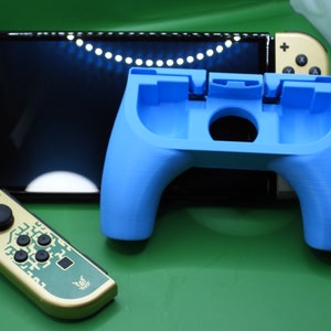 May include: A blue 3D-printed controller grip for a gaming console, with a black screen and gold-colored controllers in the background. The grip has a curved design with a central opening and is designed to hold the console.