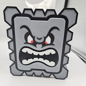 May include: A gray tissue box shaped like a Thwomp from the Super Mario video game series. The box features a black outline, red eyes, and white teeth. White tissues are visible at the top.